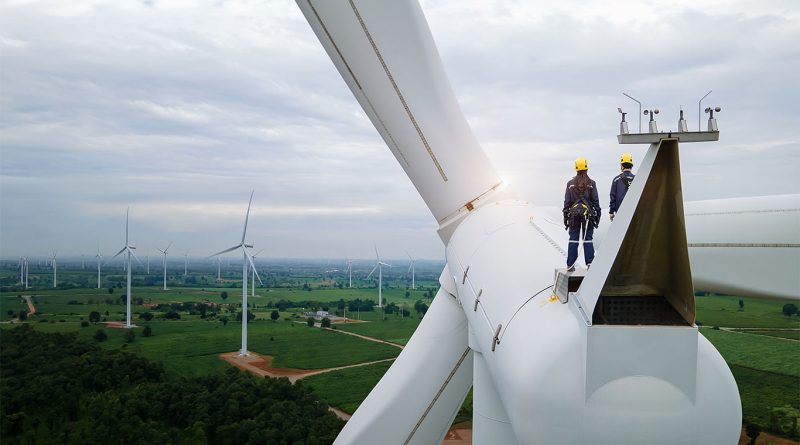 The 5 Biggest Benefits of Wind Power You Should Know Two engineers in safety gear standing atop a wind turbine nacelle, overlooking a large wind farm spread across green fields under a cloudy sky.