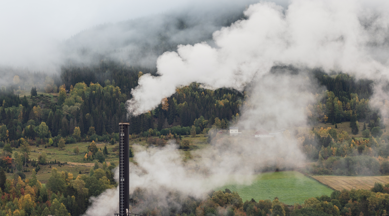 Industrial smokestack emitting white emissions over a forested landscape, symbolising the transition from fossil fuels to renewable energy