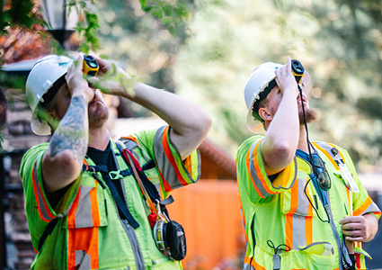 two individuals wearing white hard hats and neon green vests with orange stripes, both actively using devices to look upward