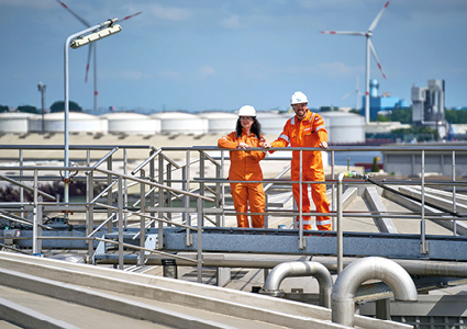 industrial workers in orange safety gear and white hard hats standing on a metal gantry at a National Gas facility