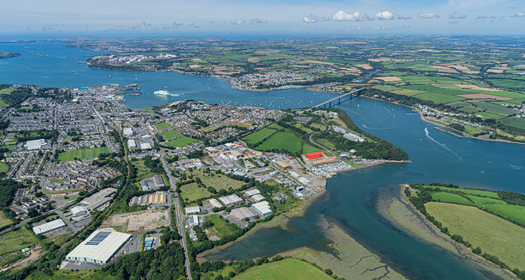 An aerial, wide-angle photograph captures a sprawling coastal landscape featuring a large waterway, a bridge, and a town surrounded by green fields under a partly cloudy blue sky