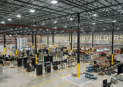 the interior of a large, high-ceiling industrial warehouse or distribution facility with exposed steel roof trusses, rows of overhead lighting, and storage racks