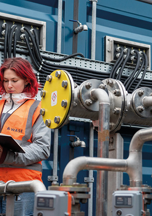 an industrial worker inspecting a complex network of metal pipes, valves, and wiring connected to a large blue unit or container
